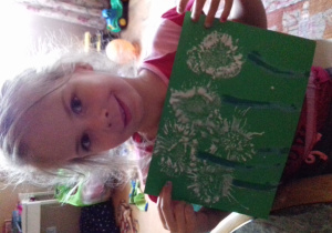 dziewczynka pokazuje swoją pracę: białe dmuchawce stemplowane pociętą rolką papieru z namalowanymi ciemnozieloną farbą łodygami