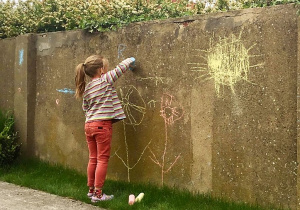 dziewczynka w kitce, w ogrodzie rysuje kredą, na betonowym płocie dmuchawce