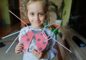 w domu uśmiechnięta dziewczynka pokazuje prace plastyczno - techniczną: biedronka wykonana z papierowego talerzyka pomalowanego na czarno, zamocowanymi do niego skrzydłami czerwonymi w czarne kropki i nogami wykonanymi ze słomek