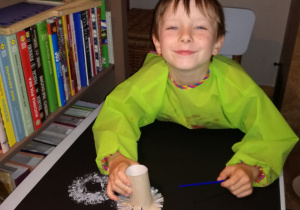 w domu, siedząc przy stole, chłopiec w zielonym fartuszku stempluje na czarnym, dużym kartonie, pociętą rolką papieru maczaną w białej farbie dmuchawce