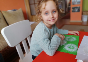 w domu, dziewczynka siedzi przy stole i stempluje białe dmuchawce na zielonej kartce