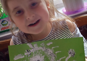 dziewczynka ze swoja pracą prac plastyczną: na jasno zielonej kartce biało czarne dmuchawce