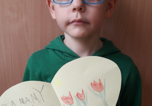 chłopiec w okularach trzyma laurkę w kształcie serca z narysowanymi: kwiatami, sercami nad mamą oraz upominkiem