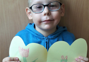 chłopiec w okularach trzyma rozłożoną laurkę, w kształcie serca z narysowanym tulipanem i prezentem