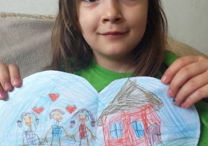 dziewczynka w ciemnych włosach trzyma przed sobą wycięte, niebieskie serce z narysowaną na nim rodziną i domem