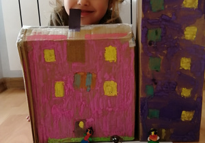 dziewczynka w jasnych kręconych włosach siedzi w pokoju za domkami z pudełek pomalowanymi kolorowymi farbami, z oknami a przed nimi stoją ludziki wykonane z plasteliny