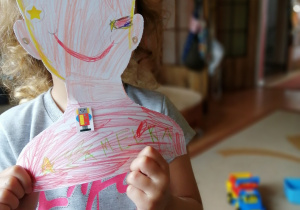 dziewczynka zasłania swoją twarz wyciętym rysunkiem twarzy, namalowanym kredkami