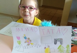 dziewczynka w okularach, w żółtej bluzce siedzi za stołem a na nim leży kartka z narysowanymi prezentami dla mamy: kwiaty, miś i góry oraz dla taty: auto, choinki i gra komputerowa