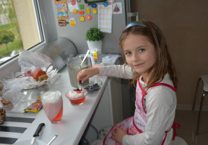 dziewczynka w domu, w kuchni siedzi przy blacie pod oknem i wkłada borówki do deseru przygotowanego z czerwonej galaretki, bitej śmietany z kawałkami truskawek