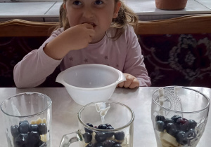 dziewczynka siedzi w domu w kuchni i je deser a przed nia, na stole stoją 3 różne szklanki z deserami z płatkami, jogurtem i borówkami