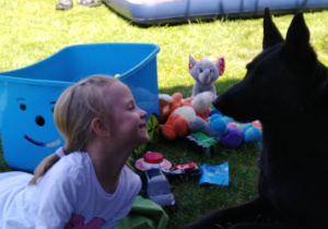 dziewczynka leży w ogrodzie, na trawie i uśmiecha się do dużego, czarnego psa