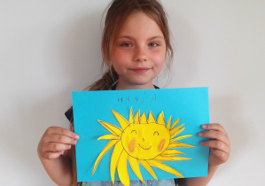 dziewczynka trzyma swoja pracę: słońce z zaokrąglonymi, trójkątnymi promieniami, zamkniętymi oczami i różowymi rumieńcami a nad nim napis Hania