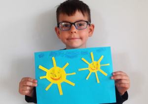 chłopiec trzyma pracę: duże i małe słoneczko podobne do siebie z prostokątnymi, równo położonymi promieniami na przeciwko siebie, rumieńcami, nosem w kształcie klamki, małymi niebieskimi oczami i czarnymi brwiami