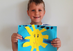 chłopiec trzyma pracę: na niebieskim tle naklejone uśmiechnięte słoneczko z: prostokątnymi promieniami, czarnymi oczkami i brwiami a nad nim granatowe chmury