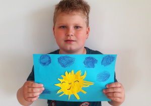 chłopiec trzyma pracę: na niebieskim tle naklejone uśmiechnięte małe słoneczko z różnej wielkości promieniami, zamkniętymi oczami i noskiem a nad nim niebieskie, puchate chmury