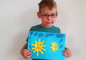 chłopiec trzyma pracę: duże i małe słoneczko podobne do siebie z prostokątnymi promieniami, okrągłymi oczami, prostokątnym nosem i różowymi rumieńcami