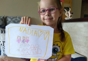dziewczynka trzyma kartkę z napisem Nadia i narysowanymi 3 koleżankami i klockami