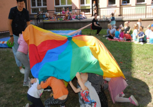 w ogrodzie podczas zabawy chustą na znak stojącej obok nauczycielki dzieci chowają sie pod chustę