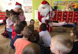 Mikołaj siedzi wśród dzieci z pomocą przebraną w czerwoną czapkę z białym pomponem