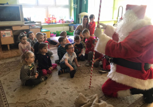 Mikołaj klęczy przed dziećmi i pomocą nauczycielki i mówi do nich