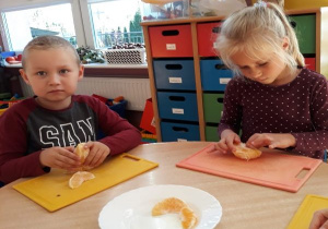 dzieci, na deskach do krojenia obierają pomarańcze a na środku stolika jest biały talerz z pomarańczami