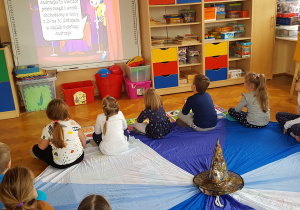 dzieci siedzą przed tablicą multimedialną i oglądają film edukacyjny o zwyczajach andrzejkowych na tablicy multimedialnej