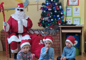 Mikołaj siedzi obok choinki z laską w dłoni a przed nim siedzą chłopcy