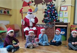 Mikołaj siedzi obok choinki z laską w dłoni a przed nim siedzą dzieci z paczkami