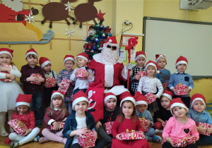 dzieci z grupy VIII w czerwonych czapkach pozują do zdjęcia z Mikołajem i prezentami