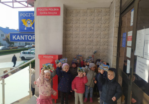 dzieci stoją przy skrzynce pocztowej pod budynkiem poczty i pokazują widokówki