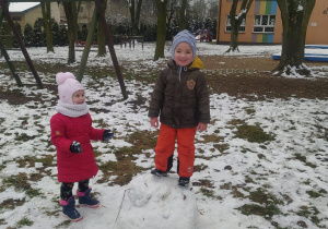 chłopiec stoi w ogrodzie na dużej kuli śniegowej a obok stoi dziewczynka