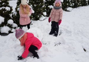 dwie dziewczynki stoją obok wielkiej kuli śniegowej a trzecia próbuje podnieść drugą, nieco mniejszą