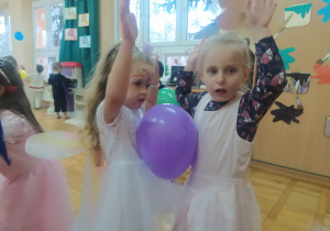 dziewczynki z rękami w górze tańczą z fioletowym balonem umieszczonym miedzy ich brzuchami