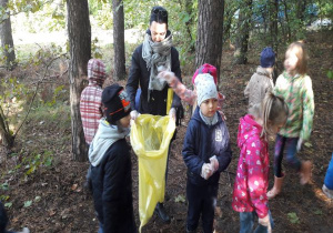 nauczycielka trzyma żółty worek a dzieci wrzucają do niego znalezione w lesie śmieci