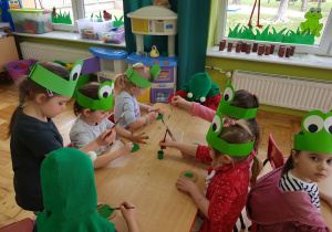 dzieci przebrane za żabki siedzą przy stolikach i malują farbami zielone kółka
