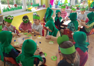 dzieci przebrane za żabki siedzą przy stolikach i kleją medale "Dzień Ziemi"