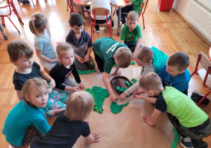 dzieci siedzą na podłodze wokół pracy grupowej - kuli ziemskiej, na szarym papierze i naklejają zieloną bibułę zwiniętą w kulki