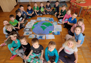 dzieci z grupy III pozują do zdjęcia siedząc na podłodze w kole a w środku wykonana Ziemia oraz indywidualne prace o Ziemi