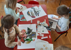 dzieci siedzą przy stoliku, przeglądają gazety, wycinają i naklejają w konturze kuli ziemskiej obrazki z nią związane