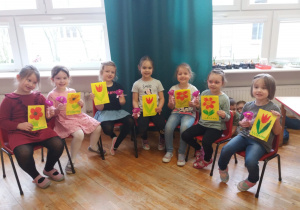 dziewczynki z grupy V siedzą na krzesełkach w półkolu i pokazują otrzymane laurki z narysowanymi przez chłopców kwiatkami