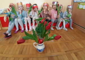 wazon z czerwonymi tulipanami stoi przed dziewczynkami z grupy III, które siedzą na czerwonej sofie w koronach, w jednej ręce trzymają tulipany a na drugiej mają założoną bransoletkę z serduszkiem