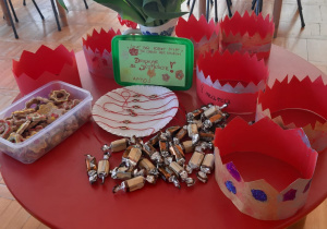 czerwony stolik a na nim korony dla dziewczynek, wazon z czerwonymi tulipanami a pod nim kartka z wierszykiem, talerzyk z ciasteczkami a drugi z bransoletkami z serduszkiem na czerwonym sznurku