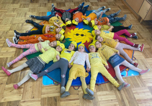 dzieci z grupy I w żółtych opaskach z naklejonymi na nich słoneczkami na głowie leżą na kolorowej chuście tworząc ze swoich ciał kształt słoneczka a w środku leży duże żółte słońce z czarnymi okularami