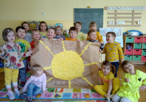 dzieci z grupy VIII pokazują swoją pracę - duże żółte słońce wyklejone kawałkami żółtego papieru, na dużym arkuszu szarego papieru z napisem 18 III Dzień Słońca