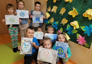 dzieci ze swoimi pracami plastycznymi o Ziemi są przed pracą zbiorową: łąka a na niej papierowe, żółte motyle i kolorowe kwiaty z bibuły