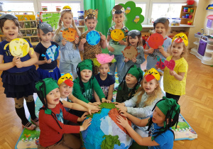 dzieci w czapkach w kształcie kwiatów, drzew, ptaków zgromadzone wokół planety Ziemi wykonanej z piłki i naklejonej na niej bibuły, pokazują narysowane planety