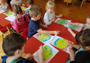 dzieci przy stolikach stemplują patyczkiem i czerwoną farbą flagę Polski jako tło do narysowanego i pomalowanego konturu mapy Polski