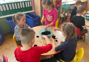 dzieci przy stolikach wsypują łyżeczkami ziemię do małych czarnych doniczek
