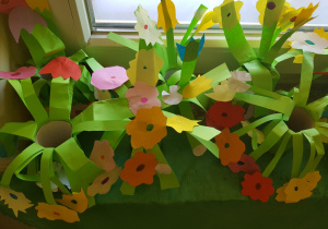 prace plastyczne - łąka z rolek papieru oklejonych zielonymi kartkami, naciętymi i zamocowanymi na końcach pasków kolorowymi kwiatami