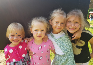 dziewczynki z kolorowymi piegami na twarzach ściskają się i uśmiechają, stojąc przed czarną tablicą w ogrodzie